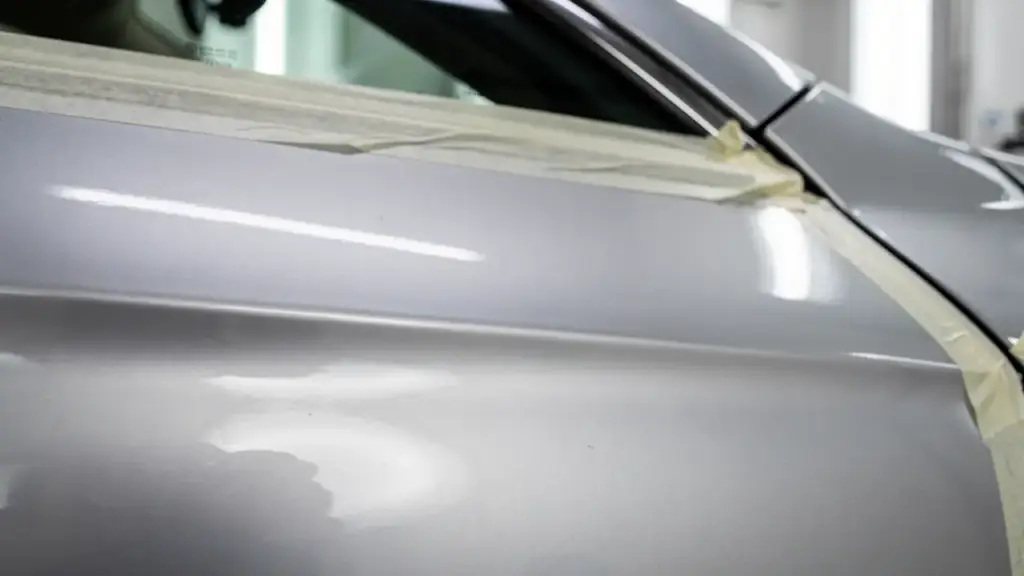 A silver car door masked with tape along the window line, the paint reflecting the booth lights